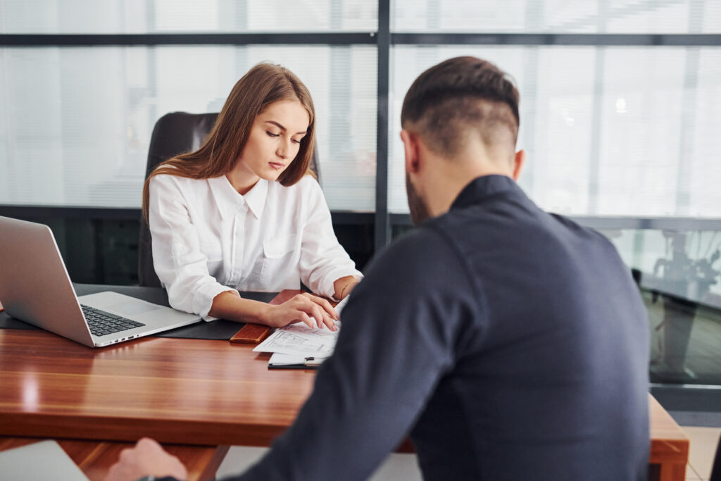 Guy sitting with documents. Woman and man in formal clothes working together indoors in the office by table.