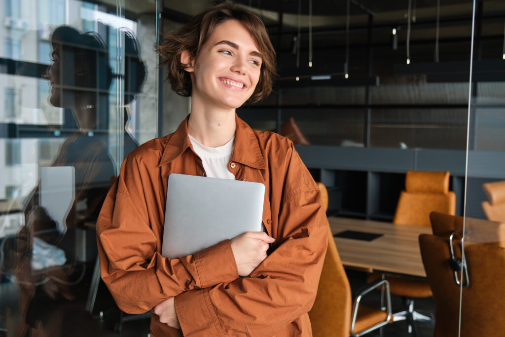 Portrait of young female manager, office worker with laptop, digital nomad programmer working at her workplace, standing with computer and smiling.