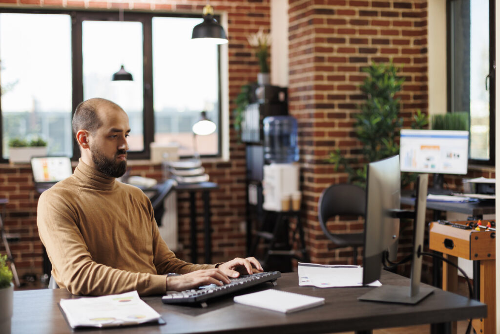 Young adult businessperson developing financial ideas and management charts. Happy smiling professional office worker looking at financial graphs while sitting at desk using work computer.